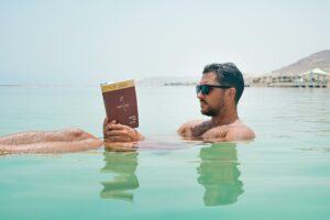 Man Wearing Sunglasses Reading Book on Body of Water